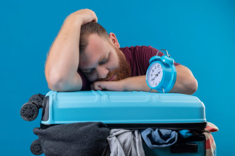 Young bearded traveler man leaning his head on suitcase full of clothes with alarm clock on it sleeping, experiencing jet lag